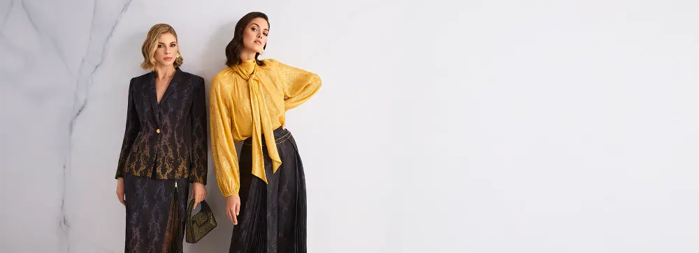 Zwei elegant gekleidete Frauenmodelle in luxuriösen Outfits mit Schlangenmuster: links ein dunkler Blazer mit passendem Midirock, rechts eine goldene Bluse mit Schleife kombiniert mit einem fließenden, dunklen Maxirock vor hellem Hintergrund.